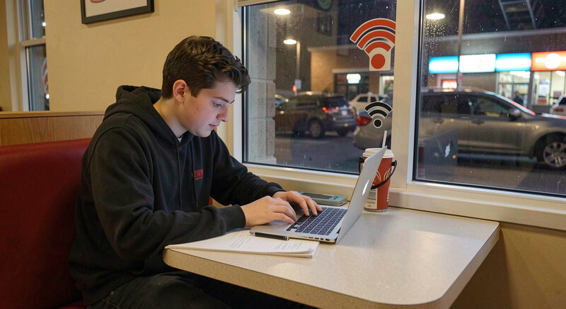 A teenager doing homework on a laptop in a fast food restaurant booth at night, near a wifi sticker on the window.