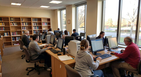 A public library computer area with several people using desktop PCs near tall windows in afternoon light.